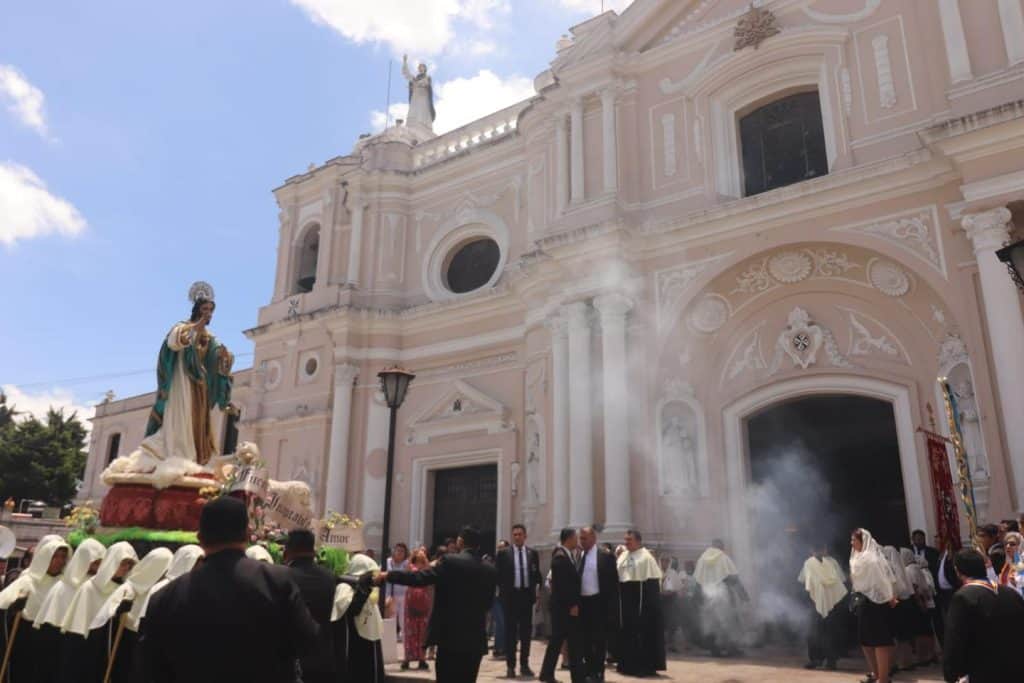 Padrinos y feligreses en una ceremonia religiosa en una iglesia barroca con detalles en color rosa y blanco, humo y una multitud participando en el acto, destacando la cultura y tradición religiosa en México.