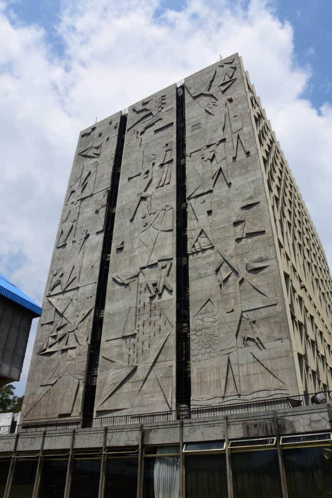 Mural oriente del Banco de Guatemala titulado Economía y Cultura de Dagoberto Vásquez Castañeda. Tiene tres paneles: A la izquiera representó la creación según la cosmovisión maya. En el panel del centro se enfocó en la cultura. Lo comenzó con una representación del tiempo. También muestra la vida cotidiana y la sociedad. El panel de la derecha representa la Economía. (Foto Banco de Guatemala) Si deseas conocer más detalles de los muros, clic en la imagen. – SoyMigrante.com REVISTA