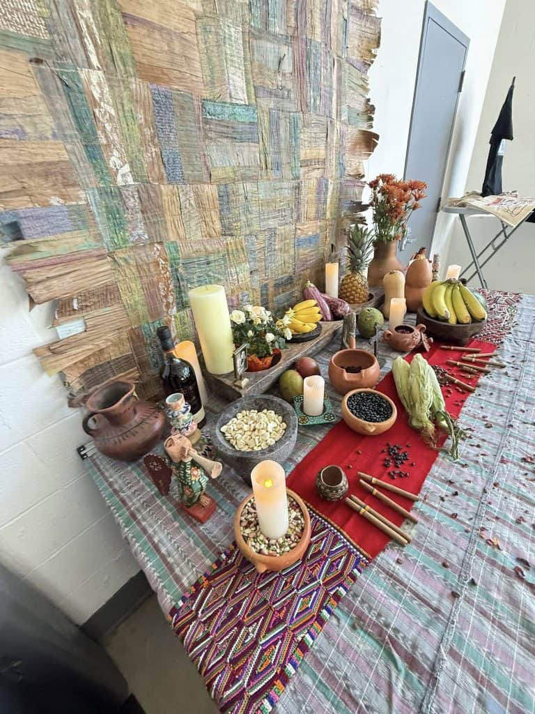 Altar en conmemoración de los seres queridos ya fallecidos, abierto en el mismo espacio de exposición de los barriletes, para que todos los asistentes pudieran meditar y depositar recuerdos. Al fondo se observa el quilt de hojas de tamal obra de la artista guatemalteca Magda León. – SoyMigrante.com REVISTA
