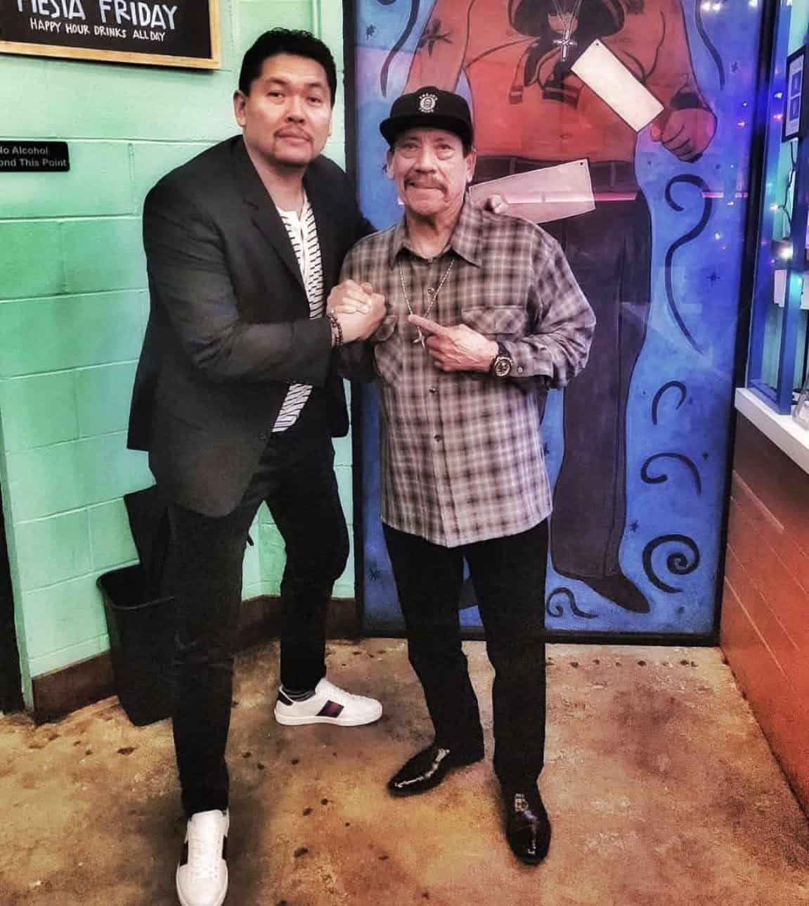 Two men pose for a photo in a colorful venue; one in a black blazer and white sneakers shakes hands with an older man in a plaid shirt and cap, pointing at him with a mural behind them.