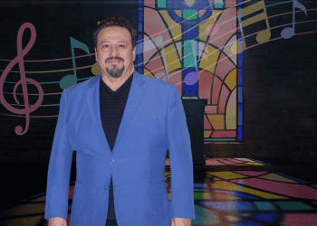 Man in a blue blazer posing in front of a colorful stained-glass backdrop with musical notes and stage lighting.