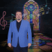 Man in a blue blazer posing in front of a colorful stained-glass backdrop with musical notes and stage lighting.