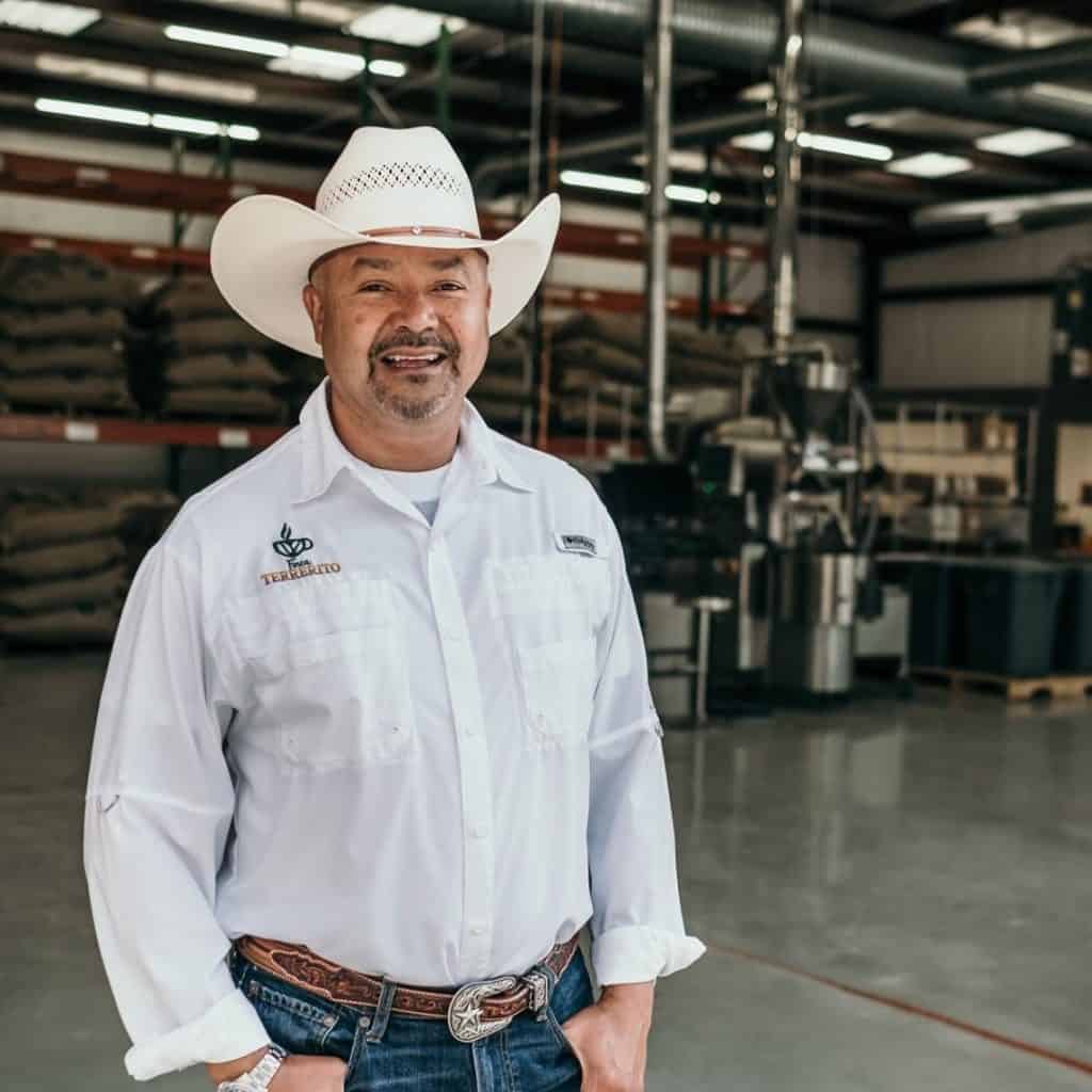 Smiling man in a white cowboy shirt and hat standing in a warehouse with industrial equipment in the background.