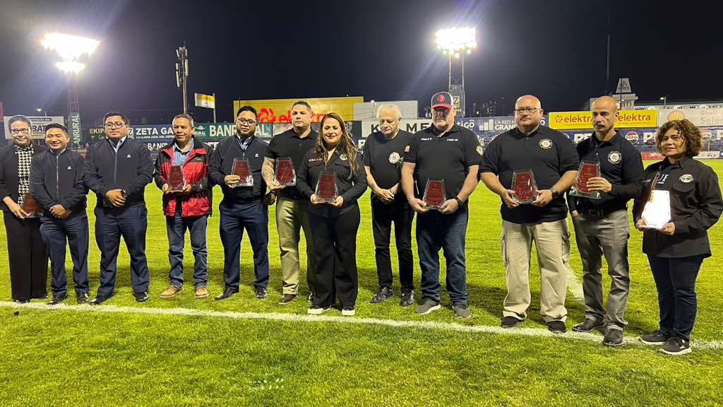 Group of people standing on a lit football field at night, each holding a red plaque or award after a ceremony.
