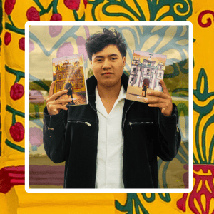 Young man in a black jacket holds up two illustrated brochures in front of a colorful mural background.