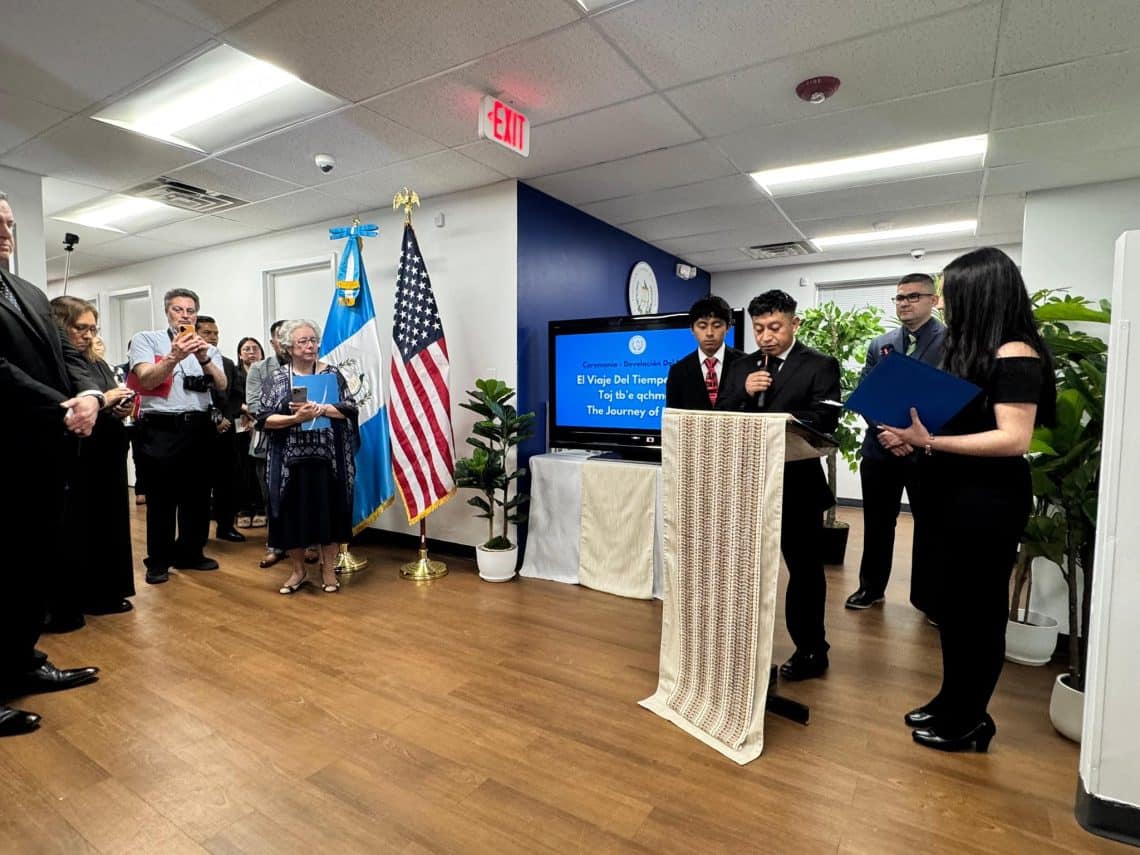 Formal ceremony in an office with flags (U.S. and another country) and a speaker at a lectern.