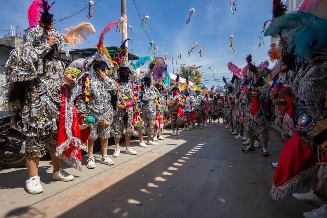 People in shimmering, fringed silver costumes with colorful headdresses line a street for a festive parade under string banners.