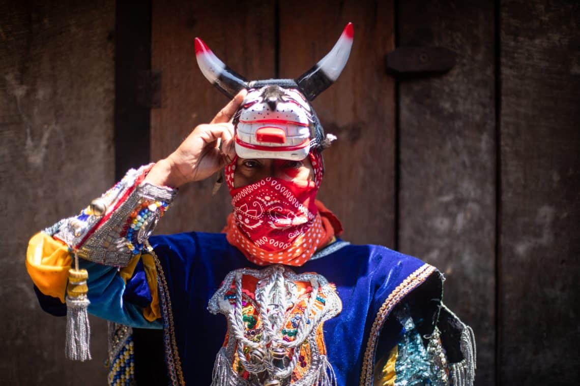 Person in a vibrant festival costume with a horned bull mask and red bandana covering the lower face, posed against a wooden background.