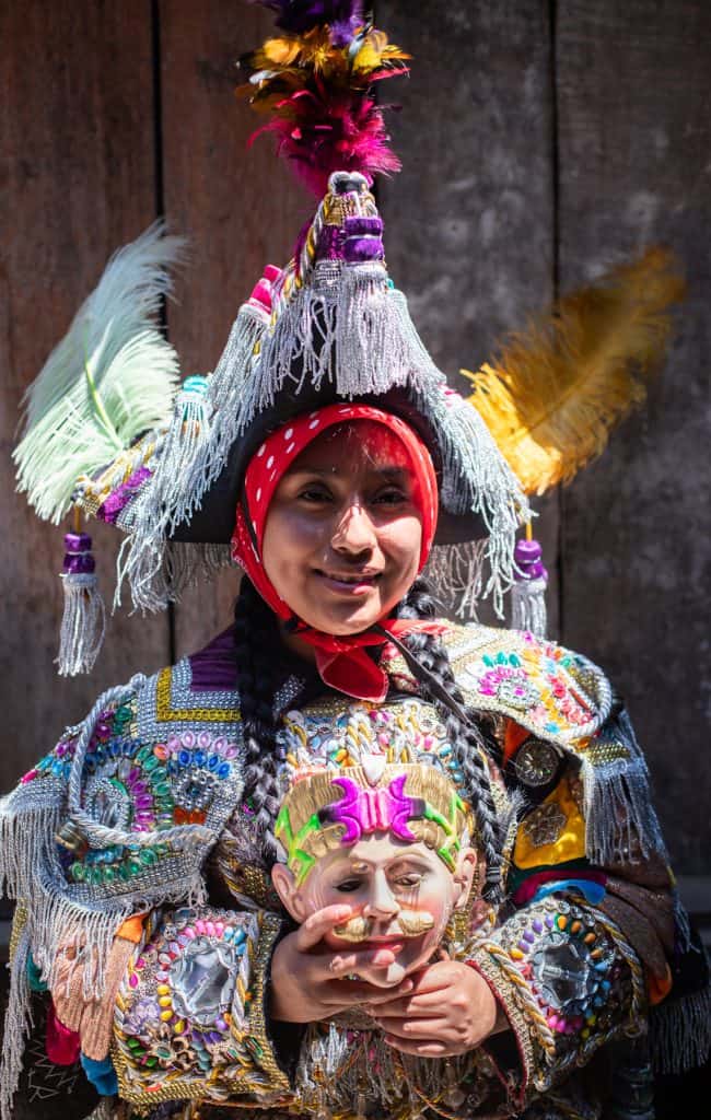 Person in a colorful, ornate costume with silver fringe, a feathered hat, and braided hair, holding a decorative mask against a wooden background.