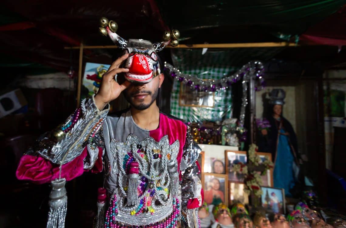Man in a colorful, ornate festive costume holding a red mask to his face in a dim market stall backdrop with glittery decorations.