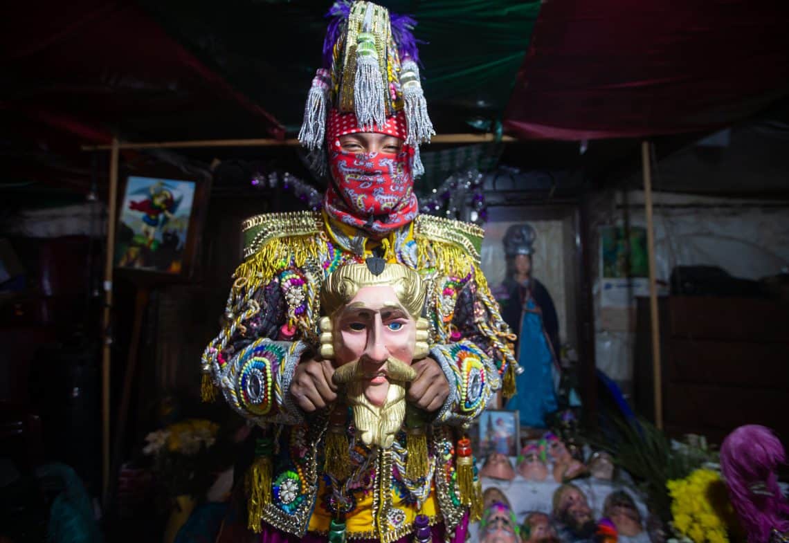 Performer in a bright, beaded carnival costume with a tall, jeweled headdress and red scarf over the face, holding a large gold mask figure in a market booth.