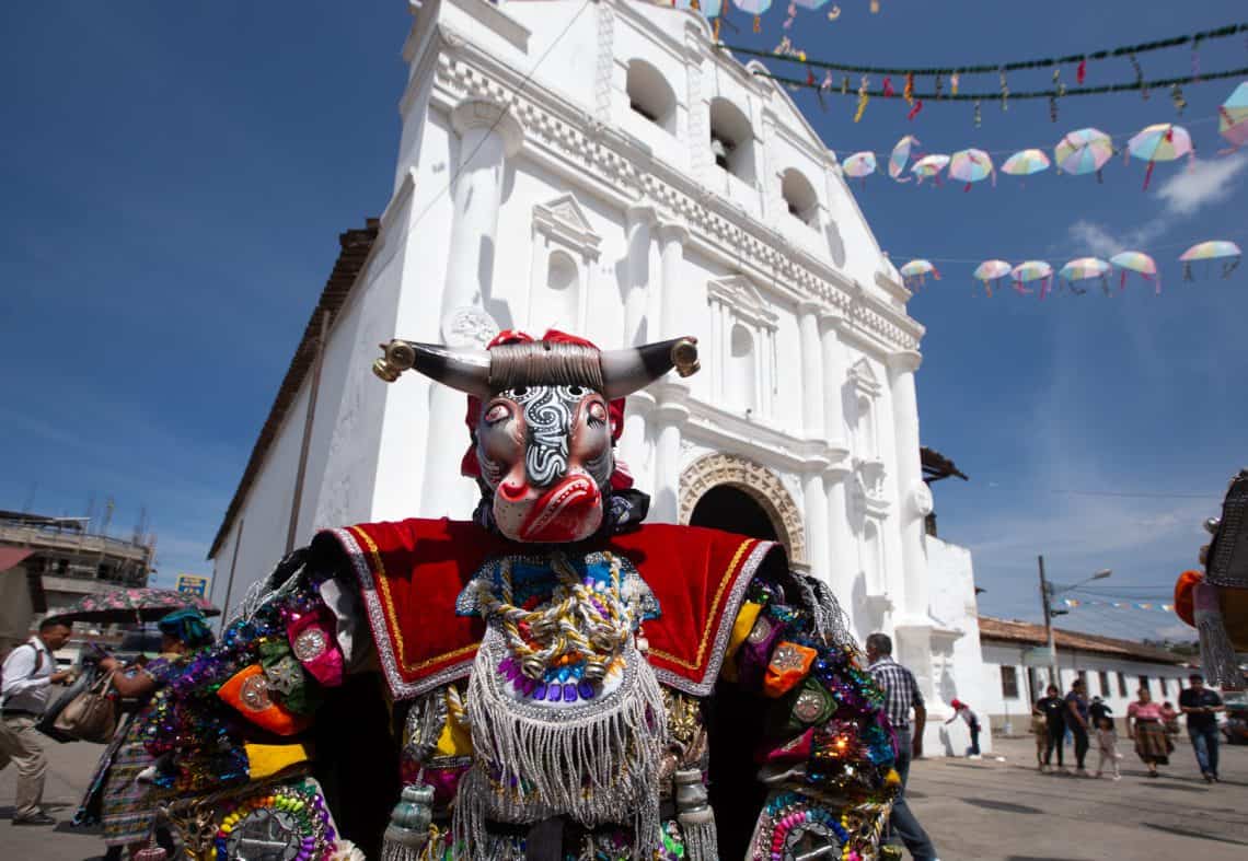 Dancer in a bright, beaded costume wearing a decorative bull mask at a street festival in front of a white church.