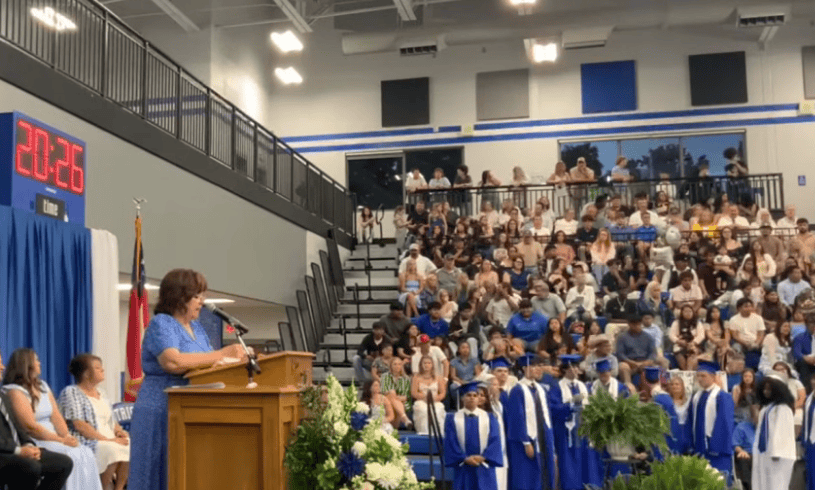 Speaker at a podium delivering a speech during a graduation ceremony in a gym, with graduates in blue gowns and a crowded audience in the bleachers.