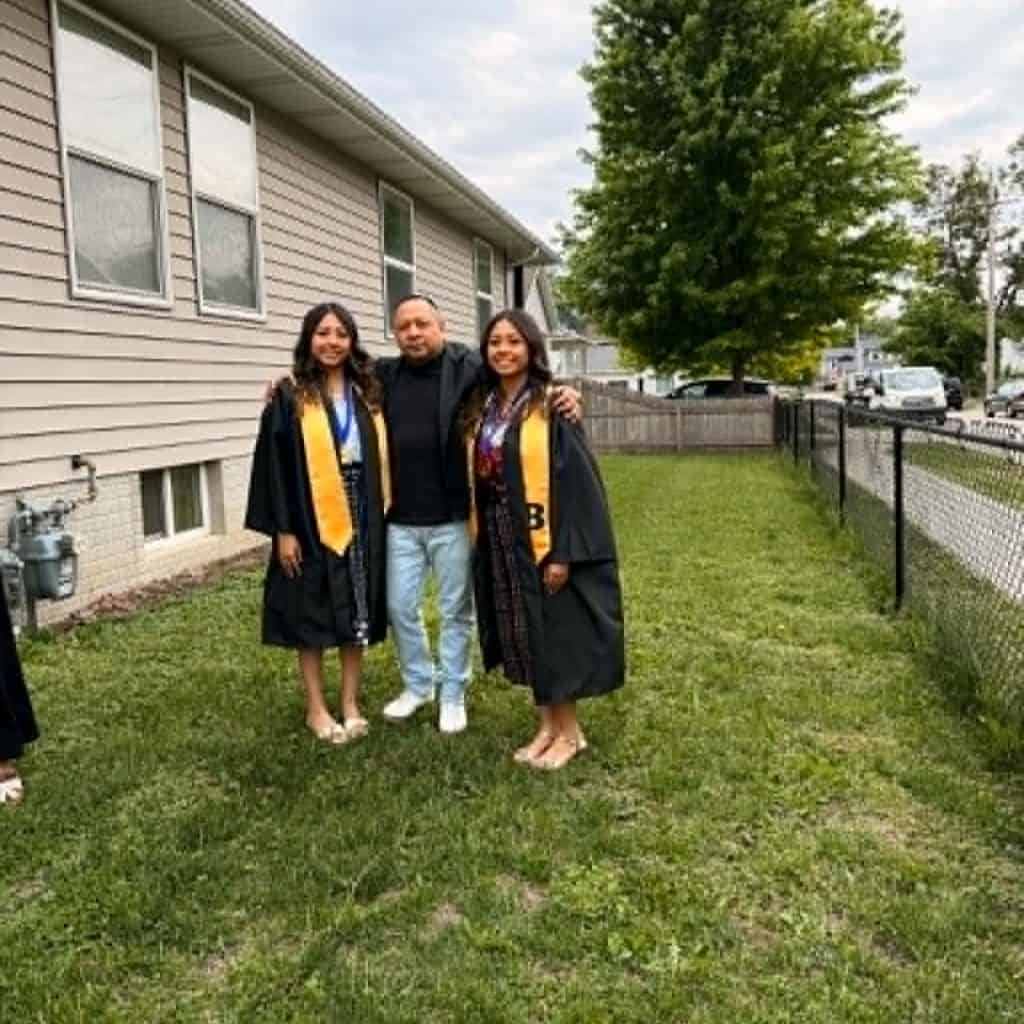 Three graduates in black gowns with gold sashes pose together on a grassy yard beside a beige house.