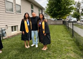 Three graduates in black gowns with gold sashes pose together on a grassy yard beside a beige house.