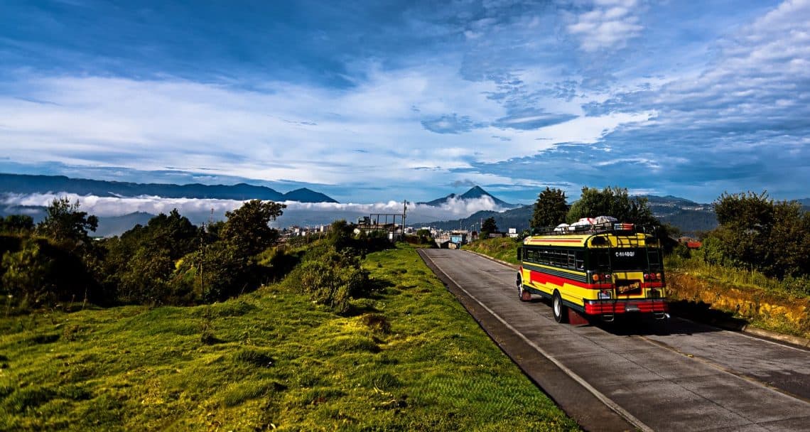 Colorful passenger bus on a rural mountain road with green fields and distant hills under a blue sky.