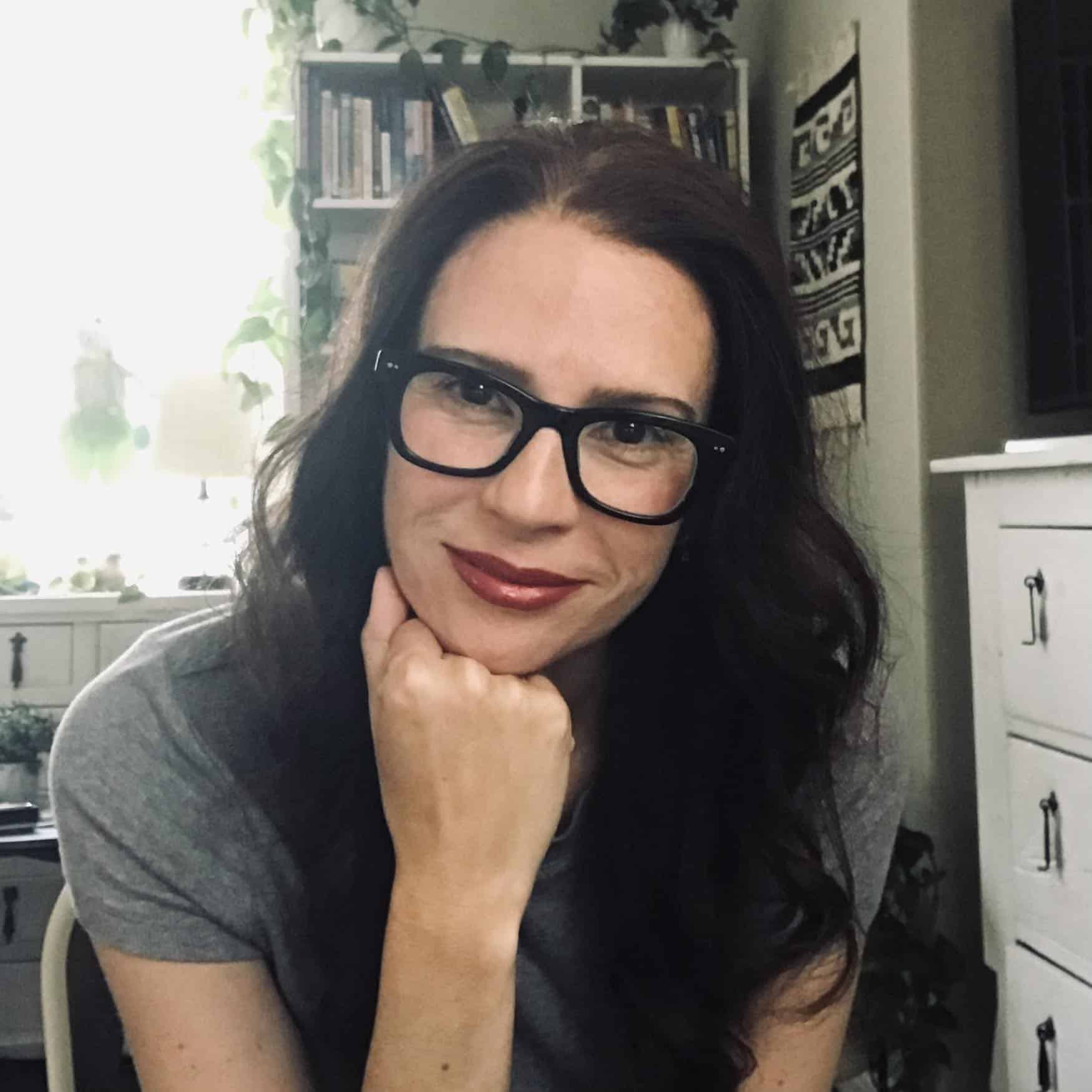 Woman with dark hair and black glasses rests her chin on her hand, smiling slightly, indoors with a bookshelf and plants in the background.