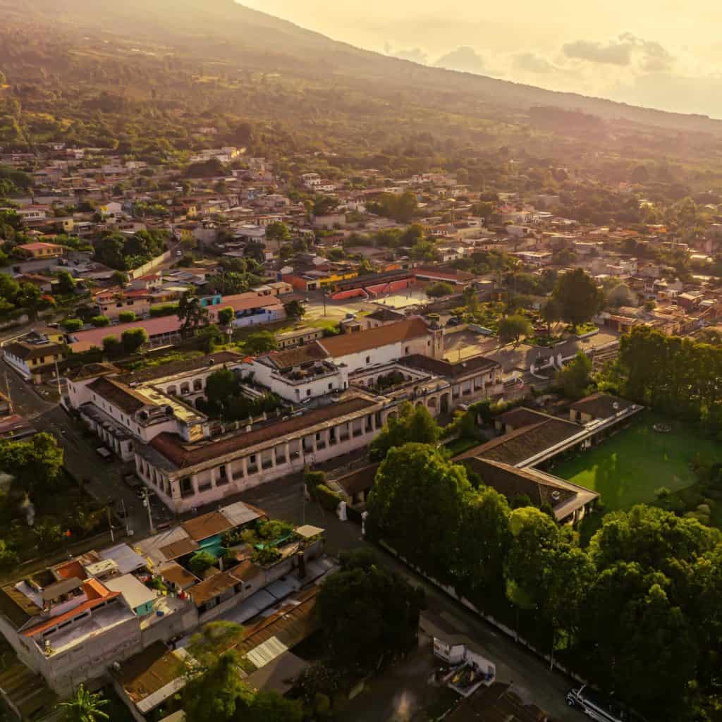 Aerial view of a hillside town at sunset, with red-tiled roofs, white buildings, and lush green trees spreading across the valley.