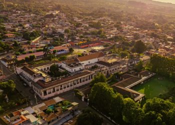 Aerial view of a hillside town at sunset, with red-tiled roofs, white buildings, and lush green trees spreading across the valley.
