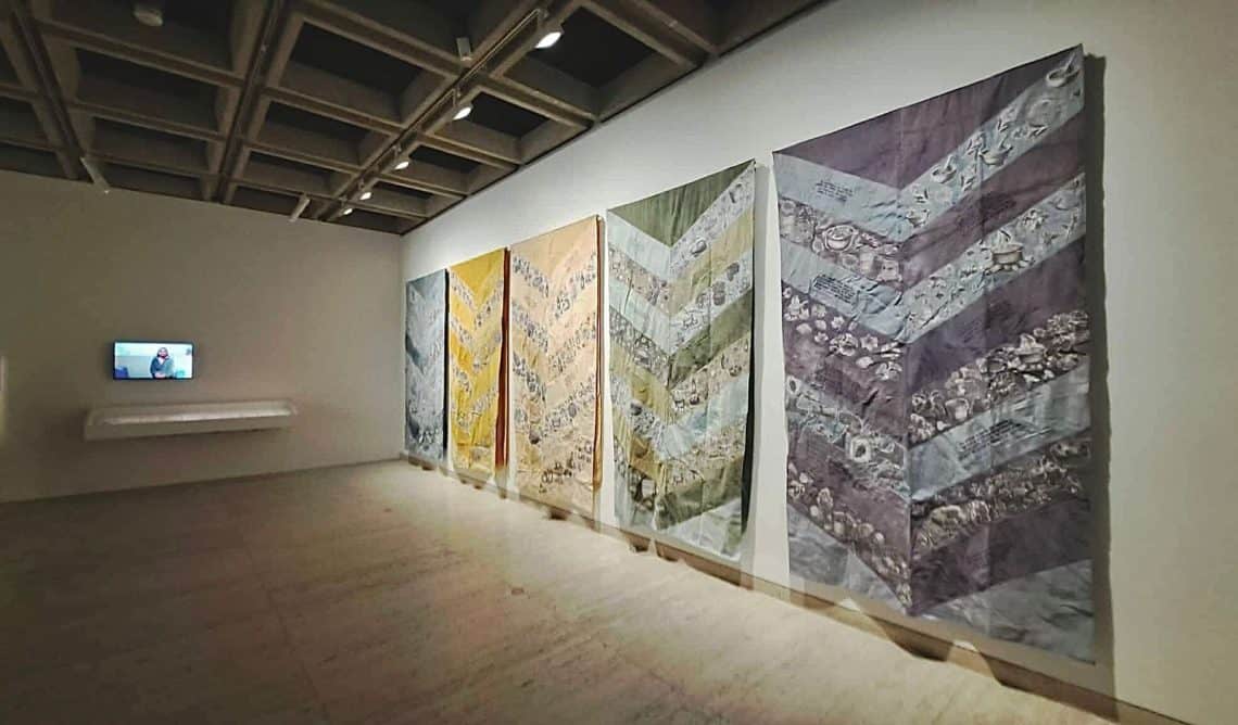 Gallery interior with five large chevron-pattern quilts on a white wall; a video monitor on the left showing a person.