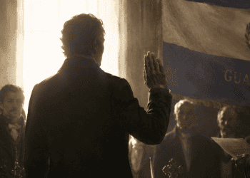 Man in a dark suit raises his hand in a formal ceremony inside a dim room, with a banner and ritual objects on a table nearby.