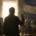 Man in a dark suit raises his hand in a formal ceremony inside a dim room, with a banner and ritual objects on a table nearby.