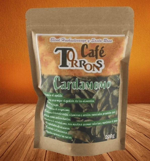 Stand-up brown resealable pouch labeled Cardamomo (cardamom) spice, placed on a wooden surface with an orange background; shows 200 g on the label.