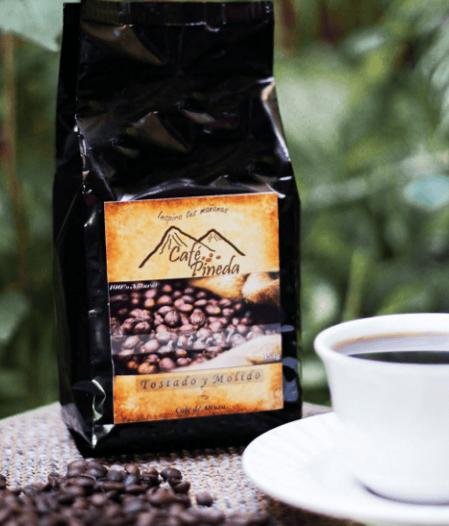 Bag of Café Pineda coffee with roasted beans spilled on a table and a white cup of coffee nearby.