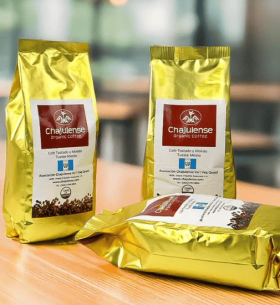 Three gold foil coffee bags with red labels reading 'Chajulense organic coffee' on a wooden table; one bag lies flat while two stand upright in the background.