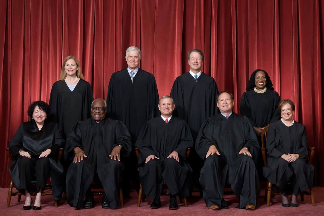 Nine judges in black academic robes pose for a formal group portrait in front of red stage curtains, with four standing in back and five seated in front.