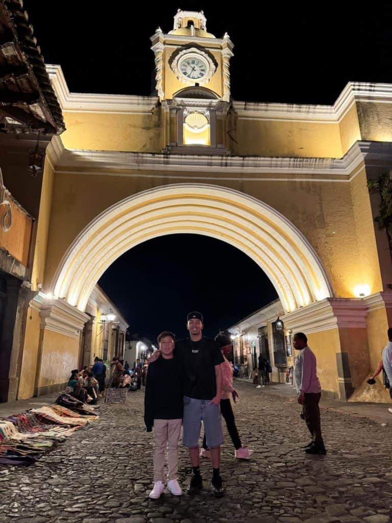 Night view of a yellow arched gateway with a clock tower, with people walking and two friends posing beneath it in the street at night.