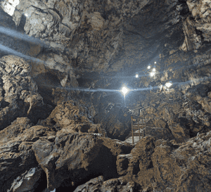 Cave interior with jagged rock walls, a metal safety railing, and bright lamps lighting a path for exploration.