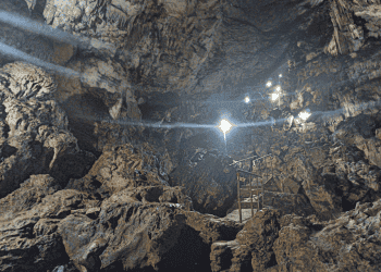 Cave interior with jagged rock walls, a metal safety railing, and bright lamps lighting a path for exploration.