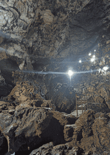 Cave interior with jagged rock walls, a metal safety railing, and bright lamps lighting a path for exploration.