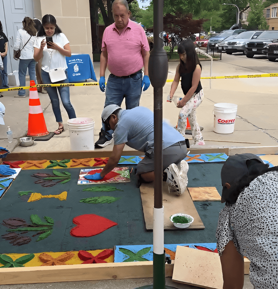 Group of people painting a large outdoor ground mural with colorful flowers and a red heart on a dark canvas.