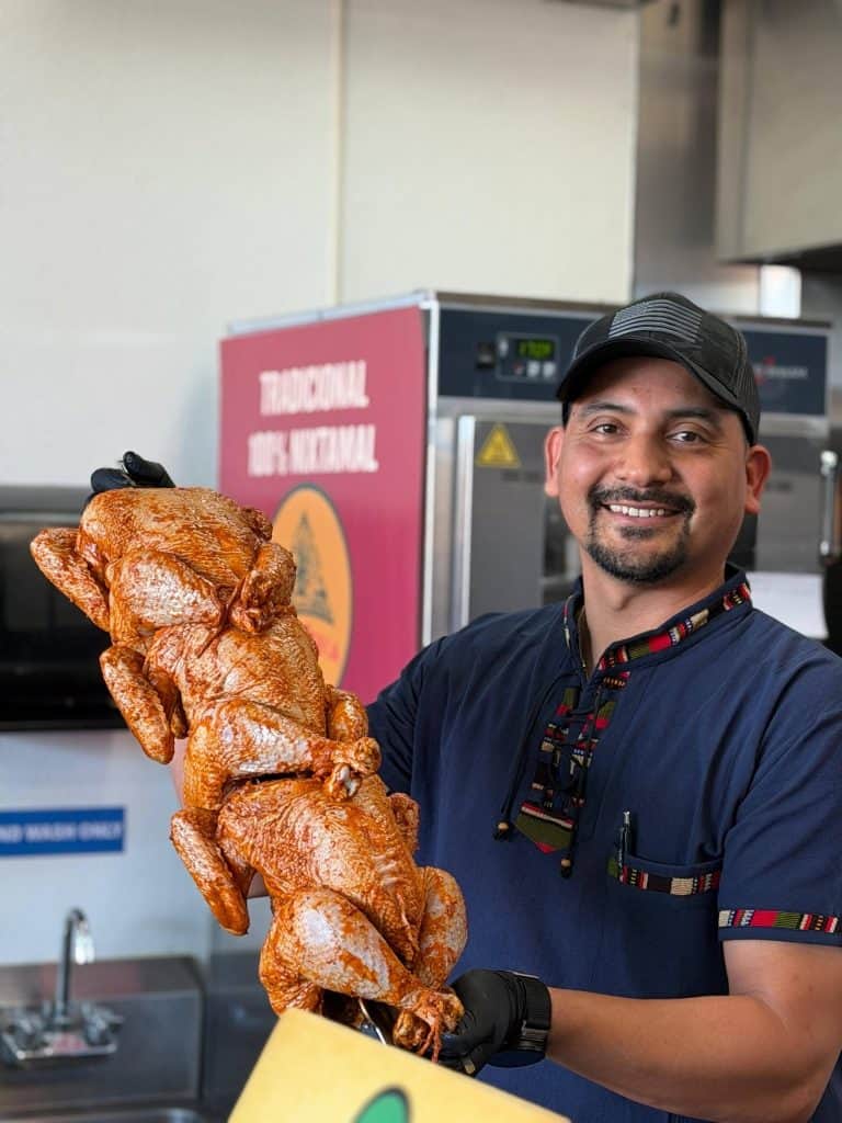 Smiling man in a cap holding a tall stack of roasted chickens in a kitchen.