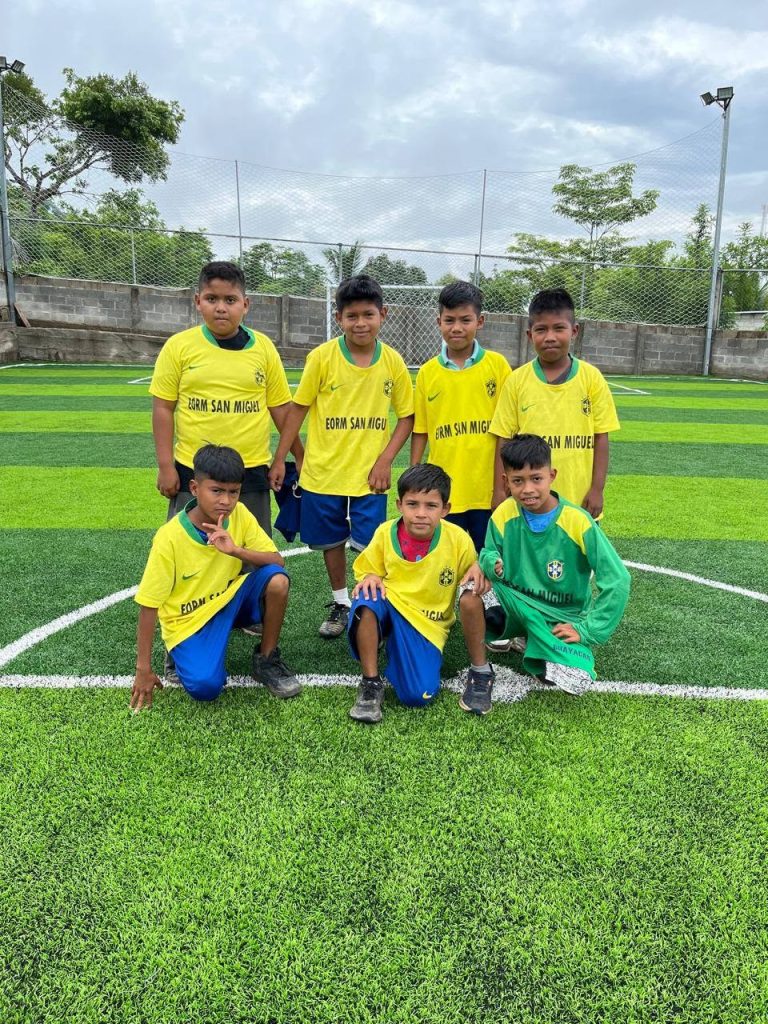 Eight young soccer players in yellow jerseys pose for a team photo on a green artificial turf field near a chain-link fence.