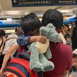 Two children hug a blue plush stuffed animal at an airport terminal, with a red backpack in the foreground and other travelers in the background.