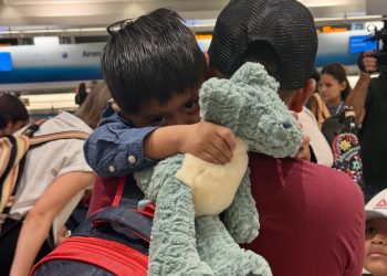 Two children hug a blue plush stuffed animal at an airport terminal, with a red backpack in the foreground and other travelers in the background.