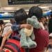 Two children hug a blue plush stuffed animal at an airport terminal, with a red backpack in the foreground and other travelers in the background.