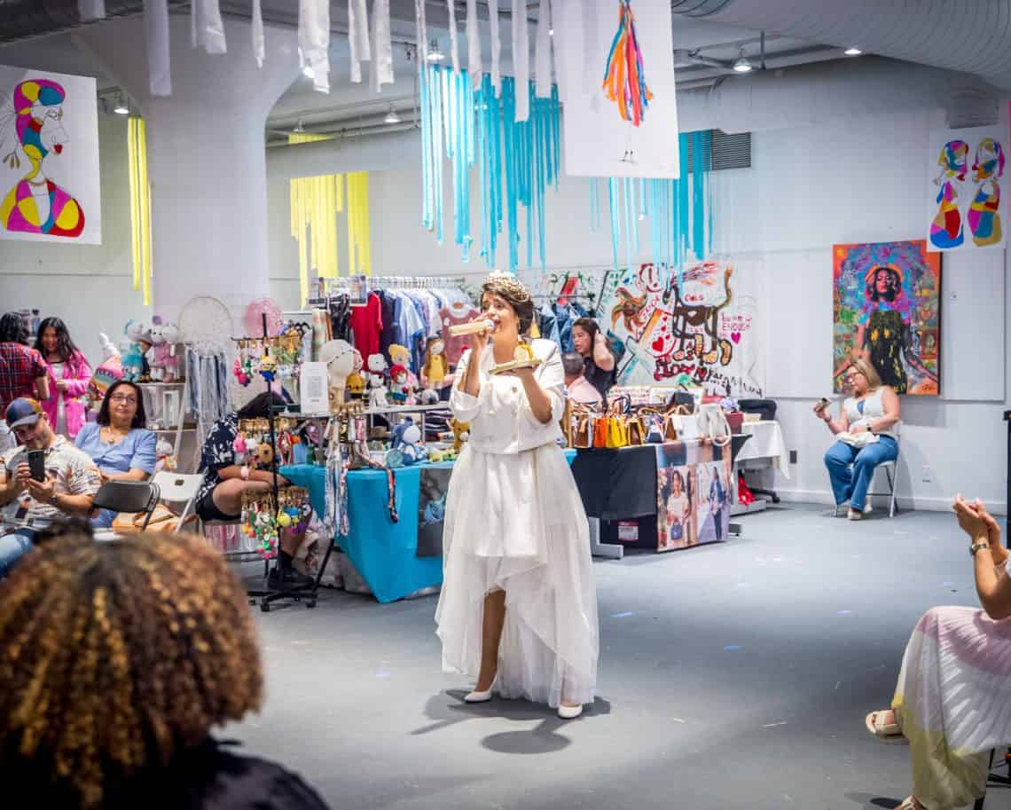 Woman in a white dress with a microphone speaking at a colorful indoor craft fair, with stalls and hanging ribbons behind her.