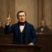 19th-century man in a navy suit raises his index finger while speaking from a wooden lectern in a courtroom setting.