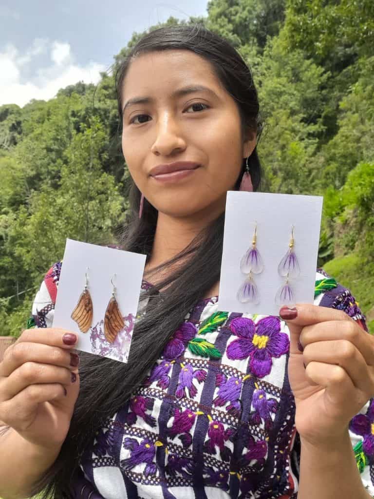 Woman outdoors presenting two earring cards: brown leaf-shaped earrings on the left and purple feather-style earrings on the right.