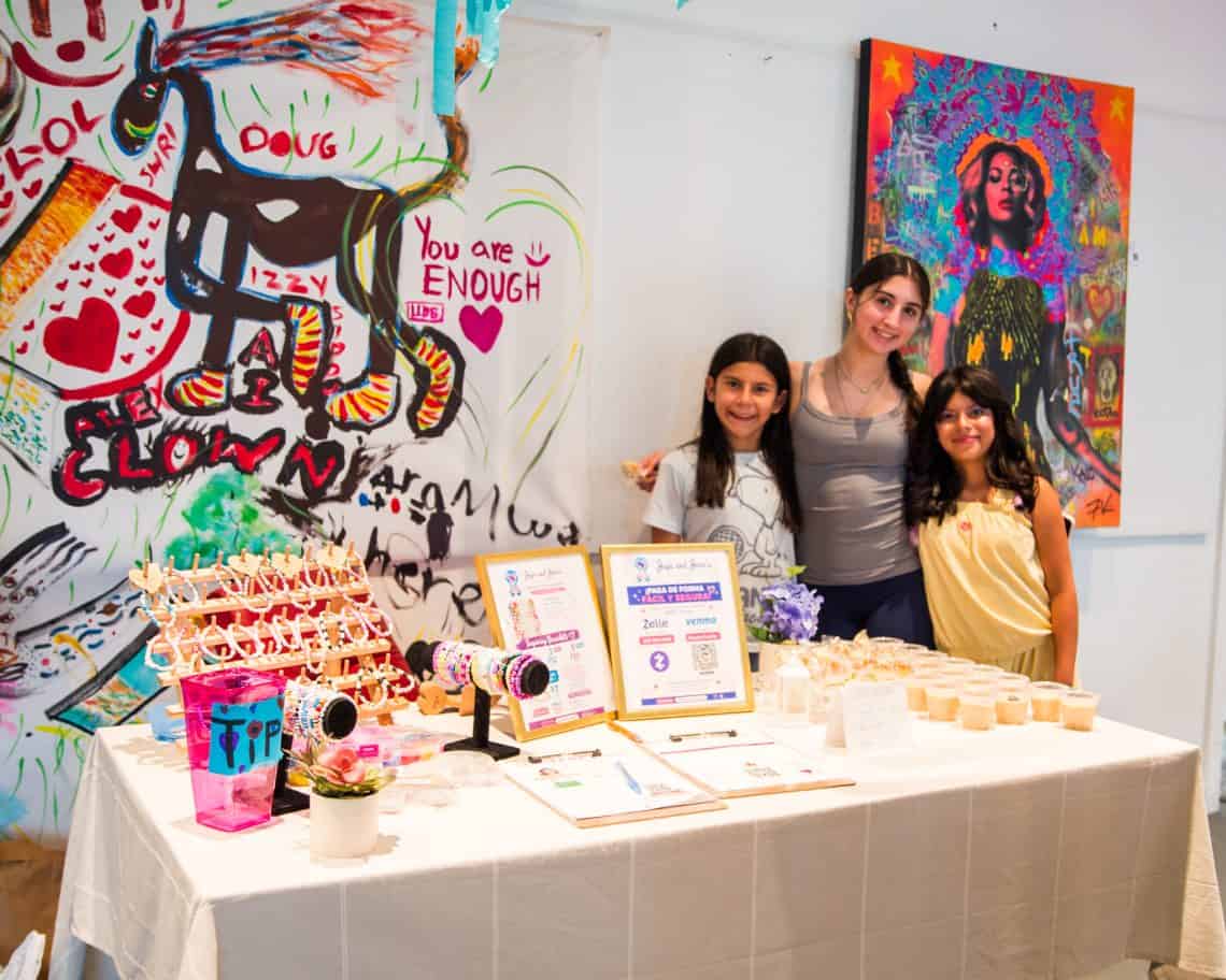 Three smiling women stand behind a table of handmade jewelry at a colorful art booth, with graffiti-style paintings in the background.