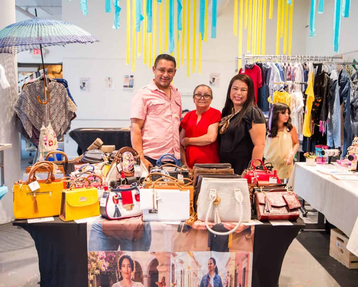 Three people stand behind a table full of colorful handbags at a clothing market stall, with racks of clothes and hanging blue and yellow streamers above.