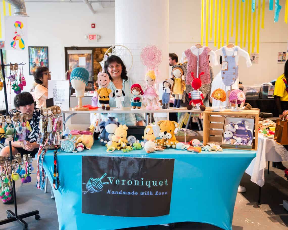 Craft market stall displaying crocheted dolls, hats, and knitted outfits, with a smiling vendor behind the table.