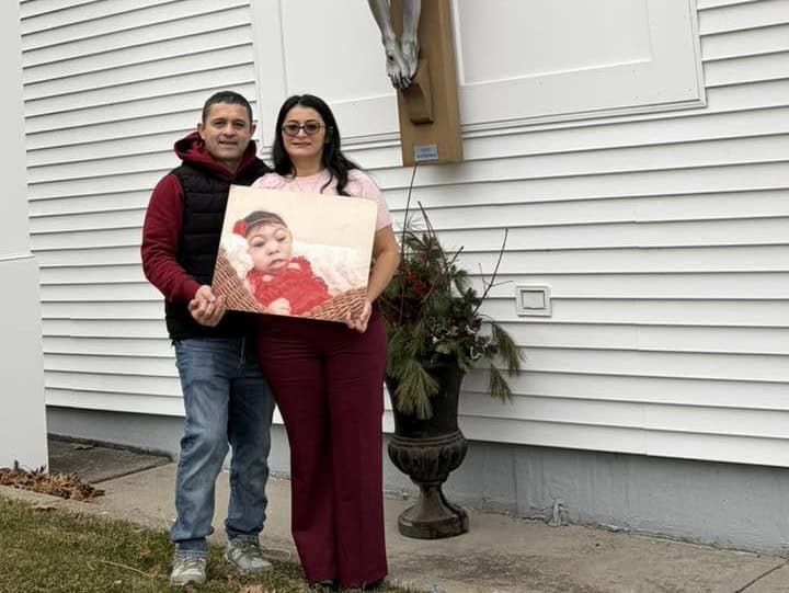 Couple stands outside a white building, holding a framed photo of a baby together.