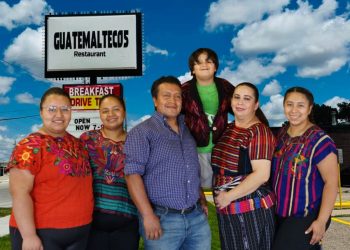Group of six people posing outside a Guatemala Meltecos Restaurant sign on a sunny day, with a child atop a man’s shoulders, all smiling.