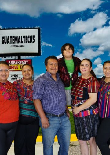 Group of six people posing outside a Guatemala Meltecos Restaurant sign on a sunny day, with a child atop a man’s shoulders, all smiling.