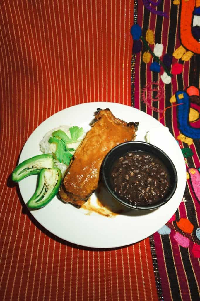 Plate with fried chicken, white rice, cilantro, cucumber slices, and a bowl of black beans on a white plate, set on a red patterned tablecloth.